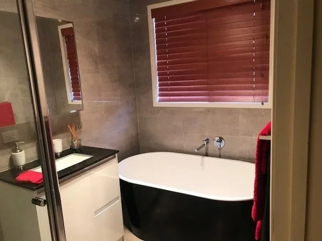 Black freestanding bath under a window with grey wall tiles