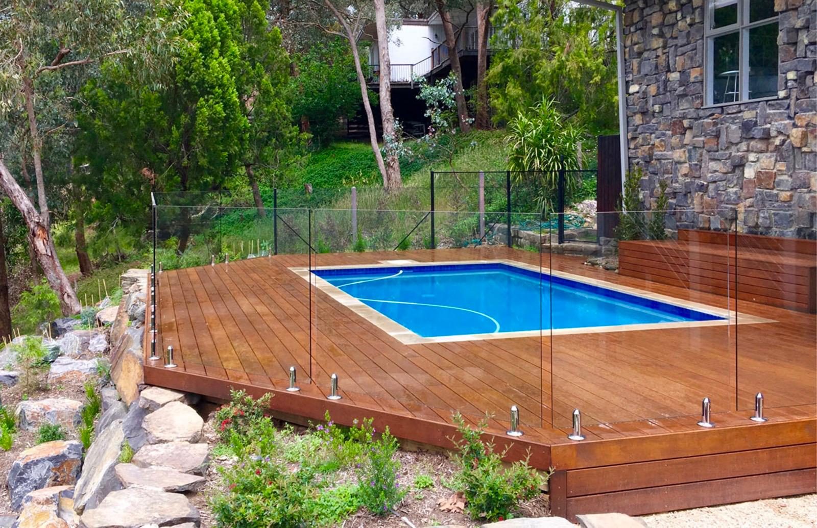Timber pool deck with glass fencing beside a stone home