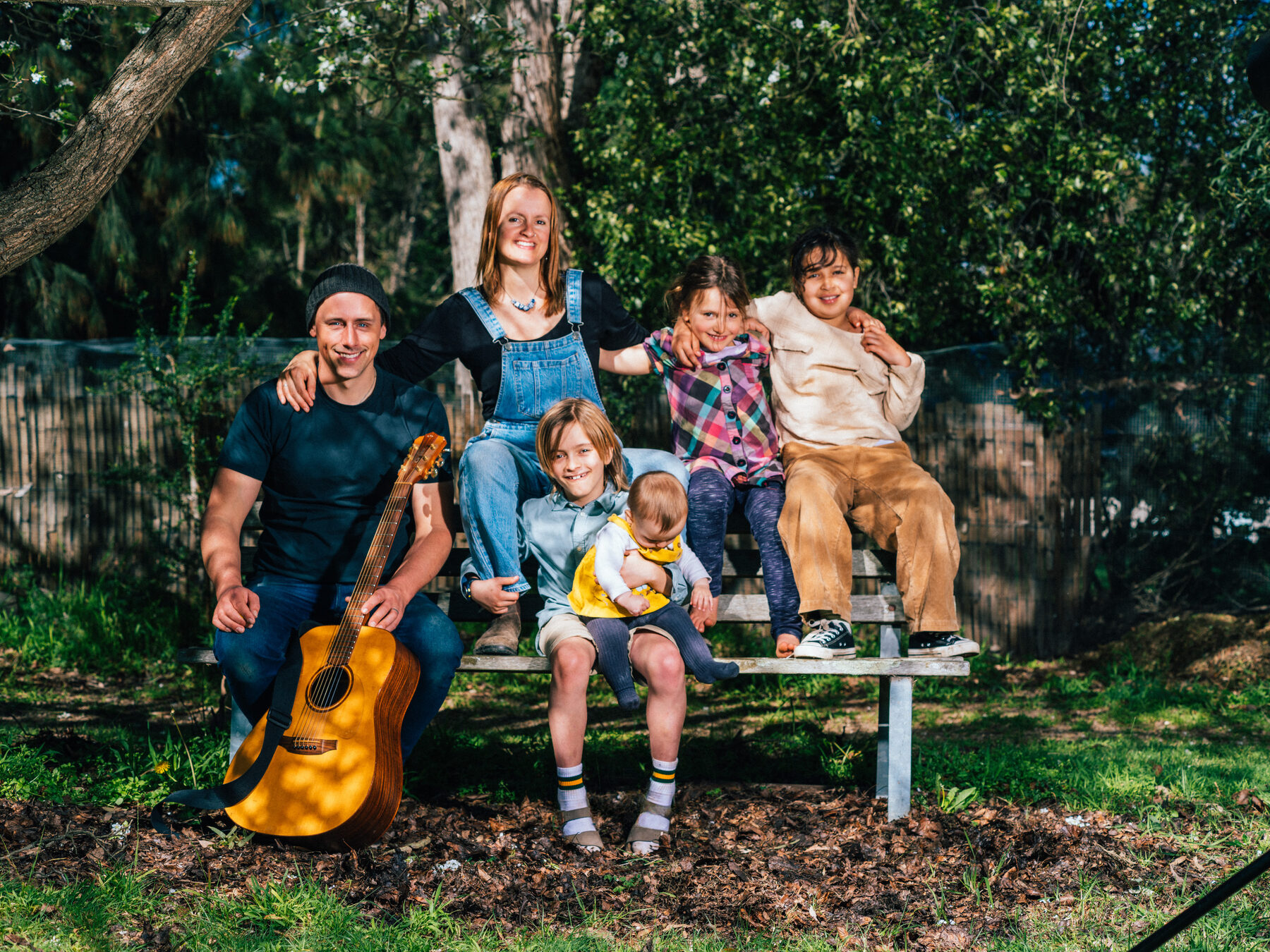 Rachel and Ollie sitting on a bench in the garden with their guitar and their children around them