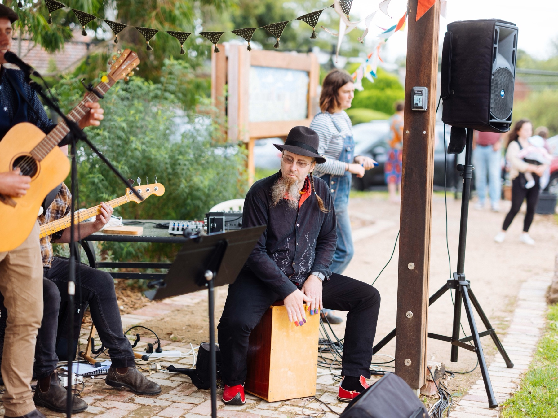 A guitarist and a cajon player performing beside the garden beds under strings of bunting
