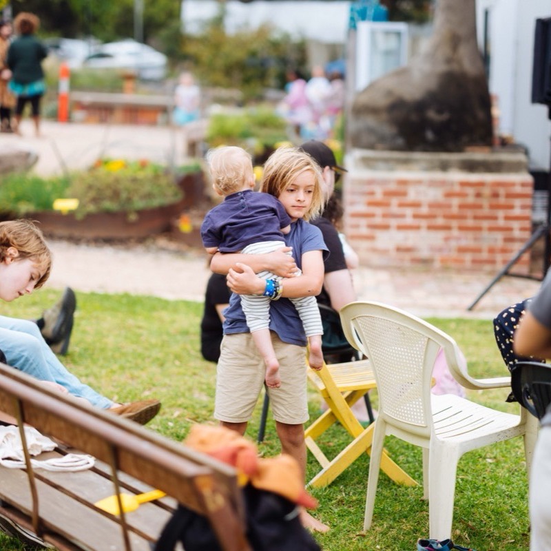 An older child carrying a small one across the grass past the garden chairs
